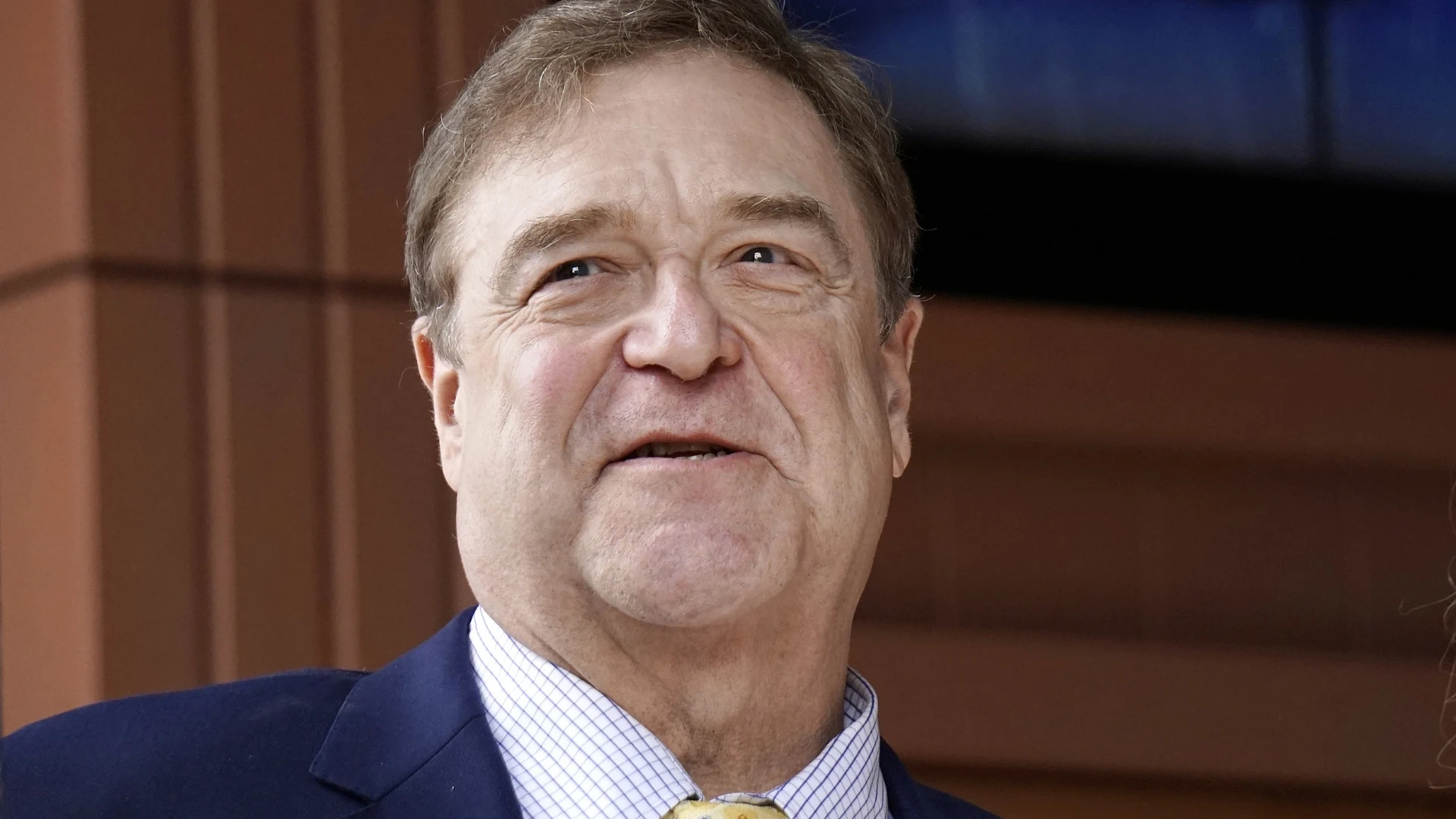 Close-up photograph of John Goodman wearing a navy blazer and a light-colored checkered shirt.