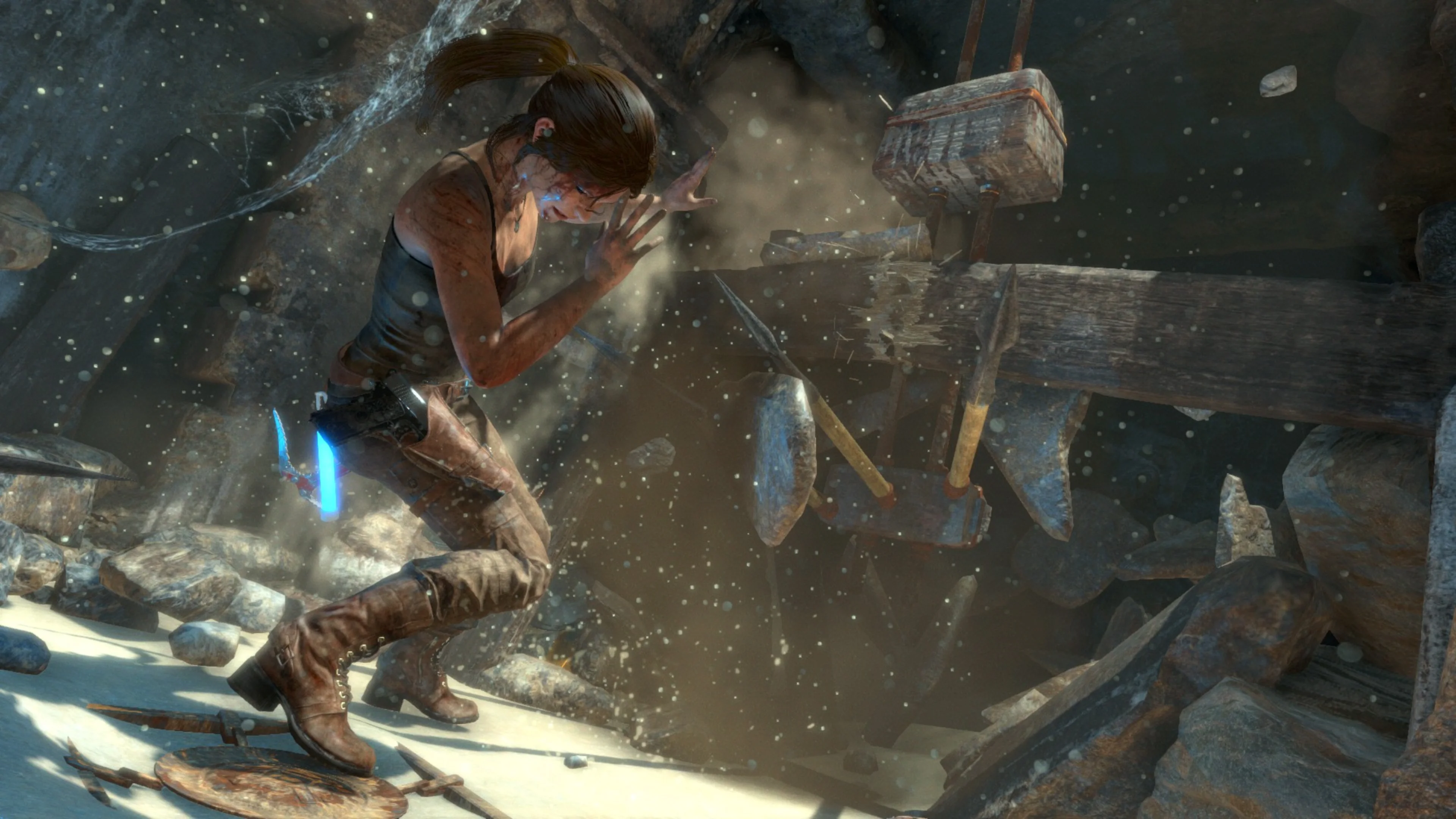 Lara Croft raises her arms to shield herself from falling debris and a wooden mechanism inside a dusty, rocky cavern.