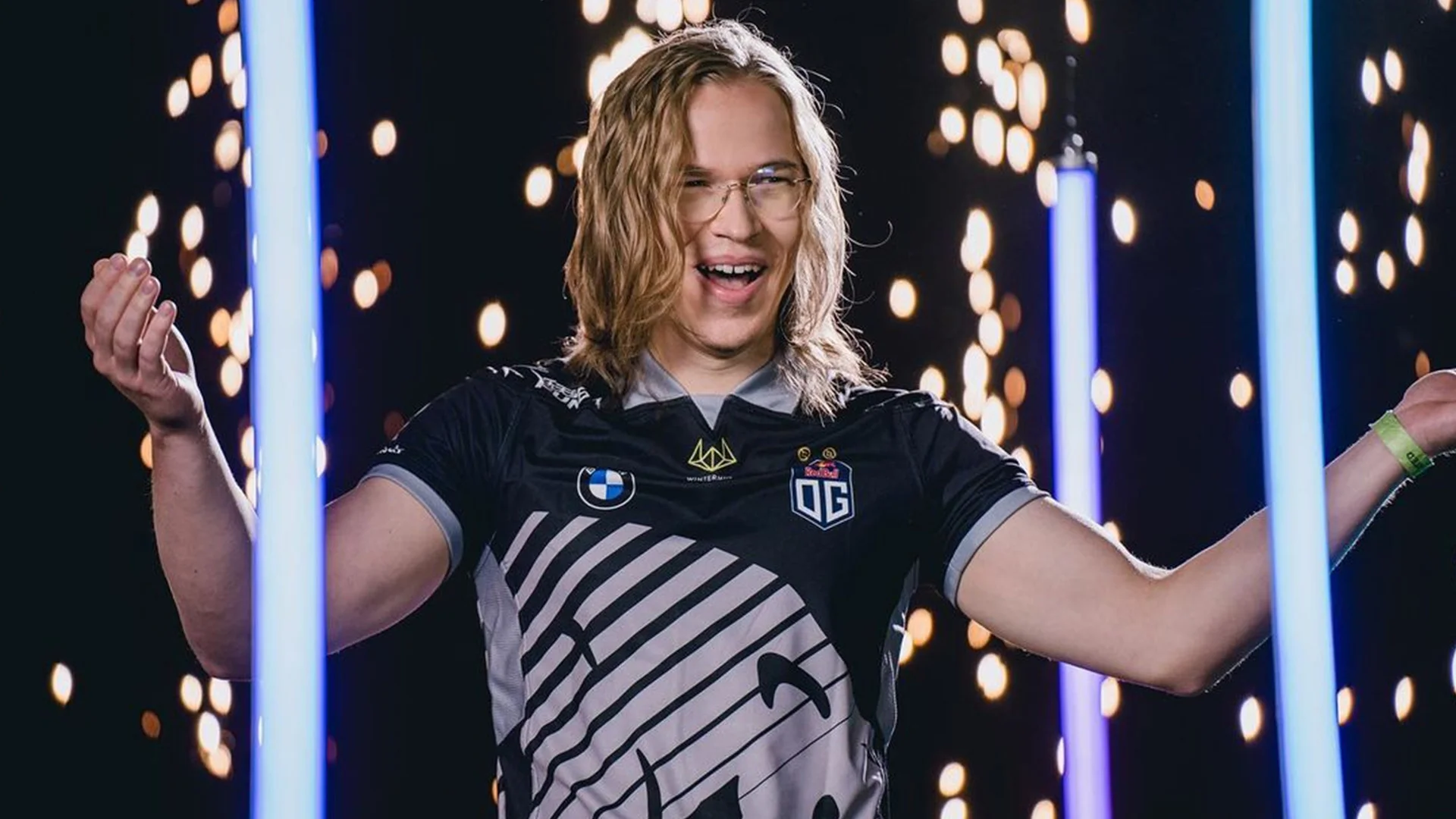 OG Dota 2 player in a black team jersey standing on stage with arms raised between blue light columns.