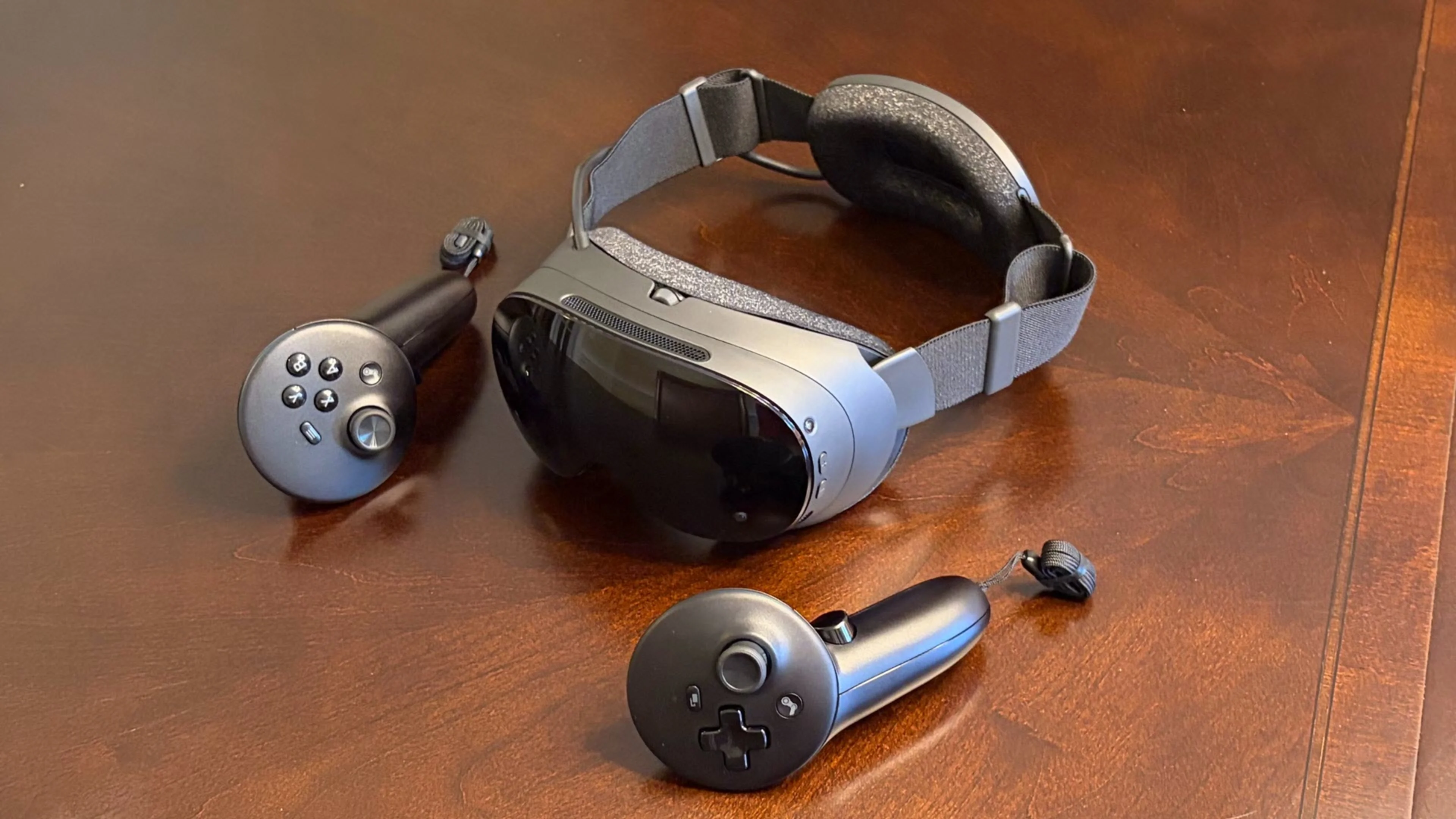 Valve Steam Frame VR headset rests on a wooden table beside two black wireless motion controllers.