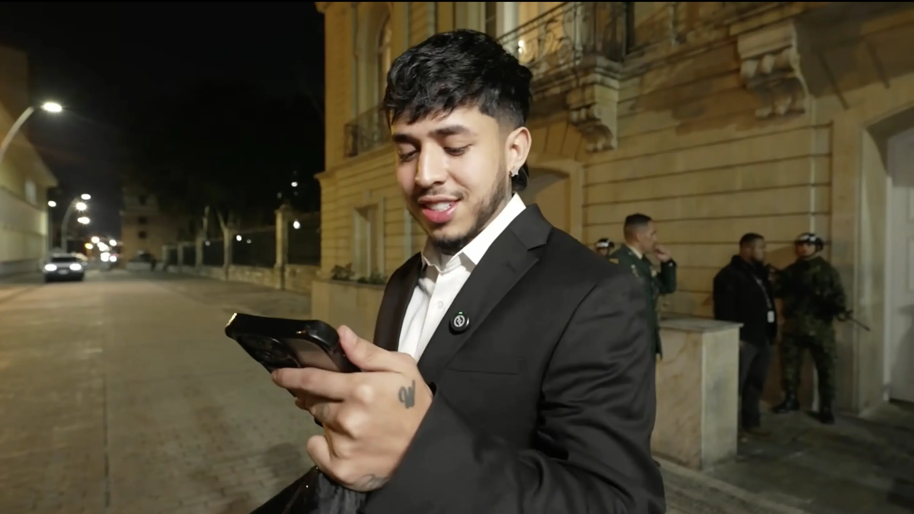 WestCOL wearing a black suit and looking at his phone outside a venue at night.