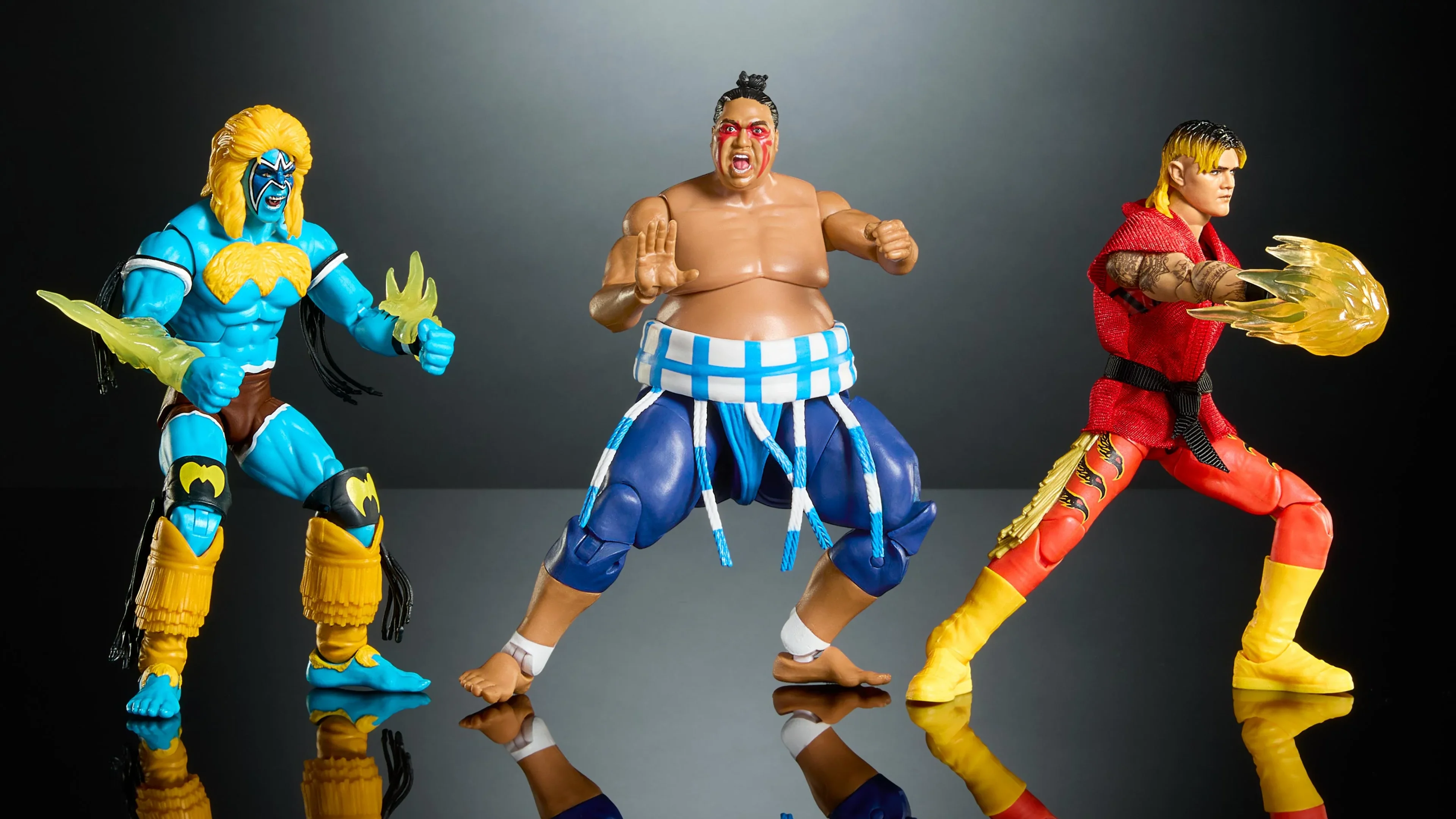 Three WWE and Street Fighter crossover action figures pose in blue, white and red wrestling outfits against a dark background.