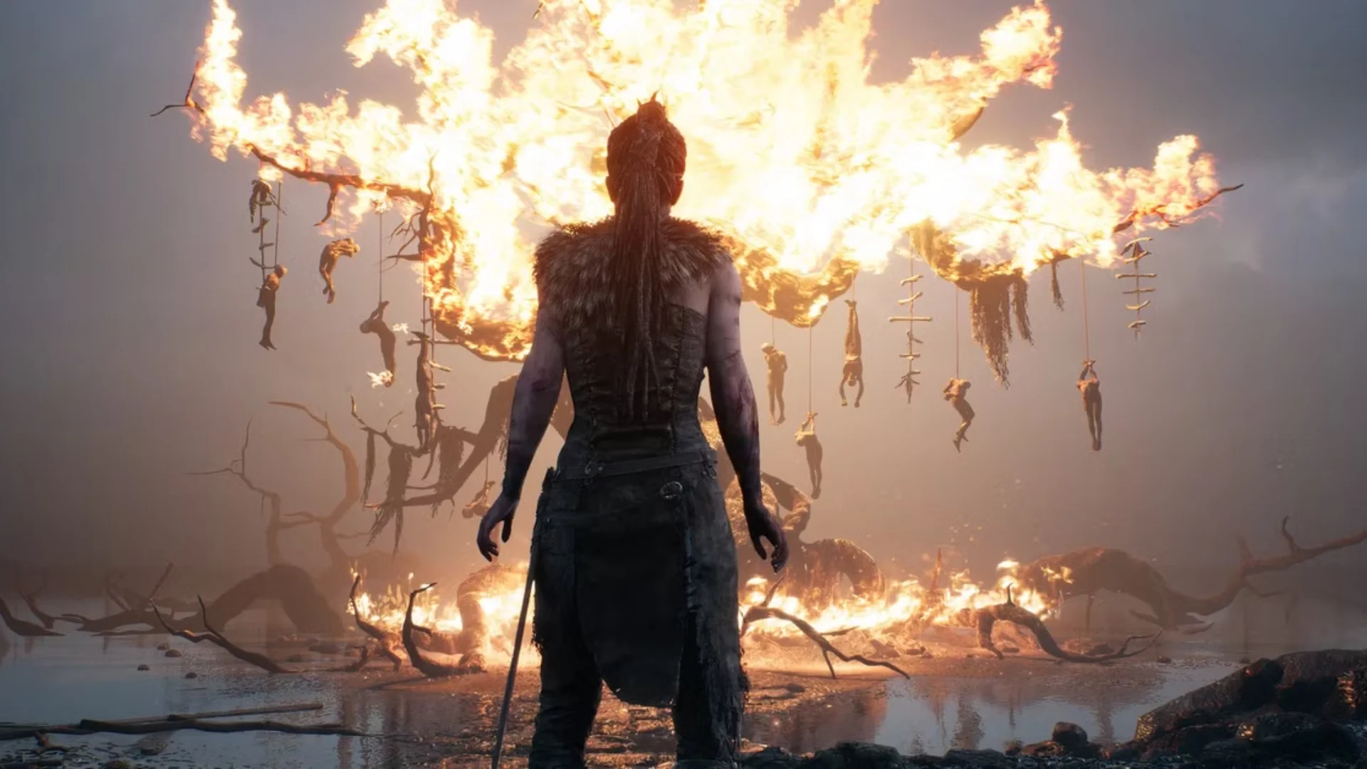 Senua stands before a massive burning tree with hanging figures in a dark Hellblade scene.