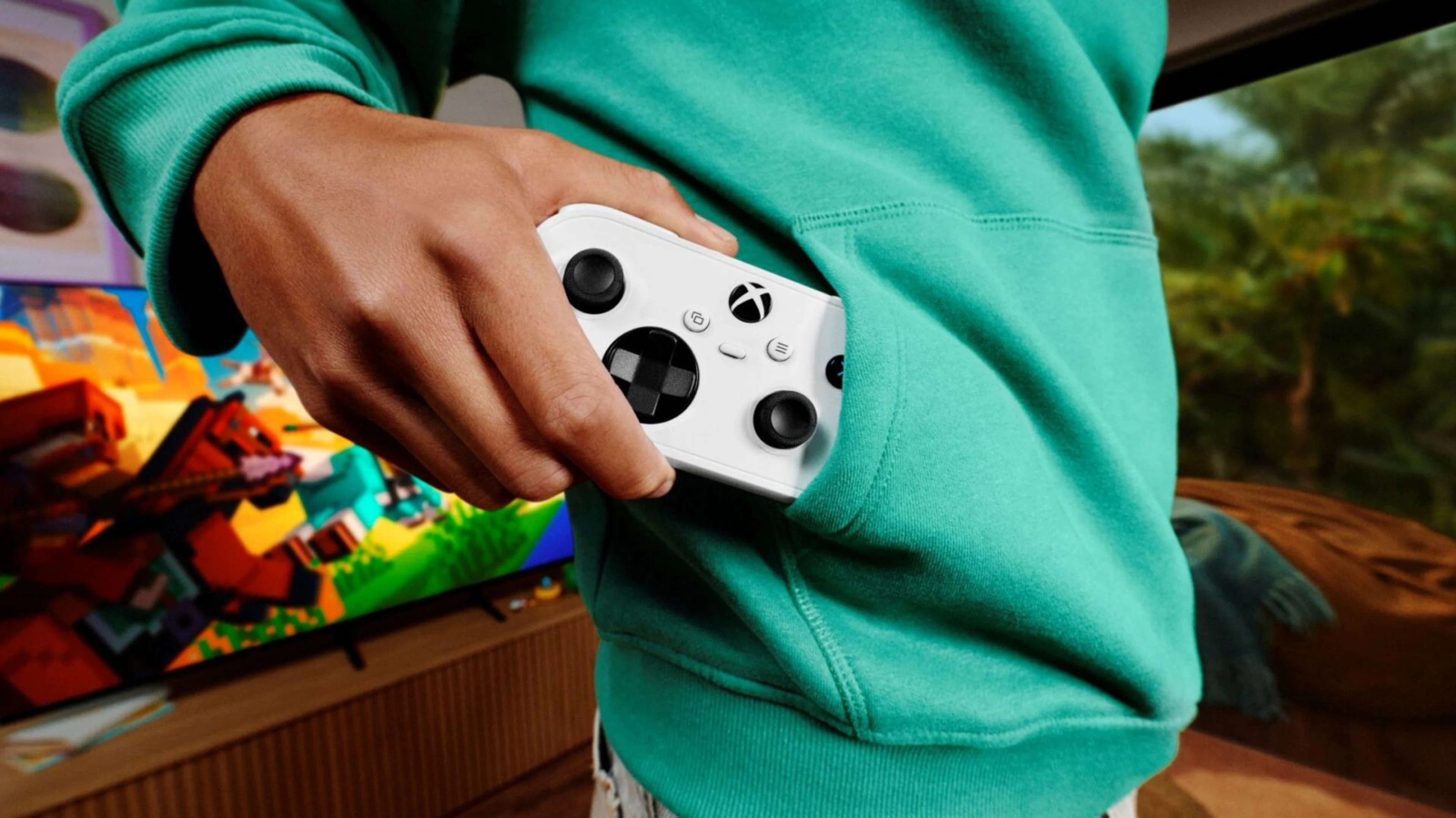 Person placing a compact white Xbox controller into the front pocket of a green hoodie, with a television in the background.